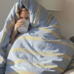 Good sleep hygiene habits of a woman sitting in a soffa covered with blue stripe sheet drinking from a cup